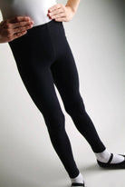 Ein Kind, das ein weißes Hemd, Danceries-Hose Danceries G30 Lexie, schwarze Leggings und weiße Socken mit schwarzen Schuhen trägt, steht mit den Händen auf den Hüften. Der Hintergrund ist schlicht und gut beleuchtet. Das Gesicht des Kindes ist nicht sichtbar.