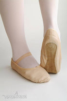 Nahaufnahme der Füße einer Balletttänzerin, die beige Ballettschuhe Liage 1050 Jump von Dance Line mit elastischen Riemen und weiße Strumpfhosen trägt. Ein Fuß steht flach auf dem Boden, während der andere angehoben ist und die Chromledersohle des Schuhs freigibt.