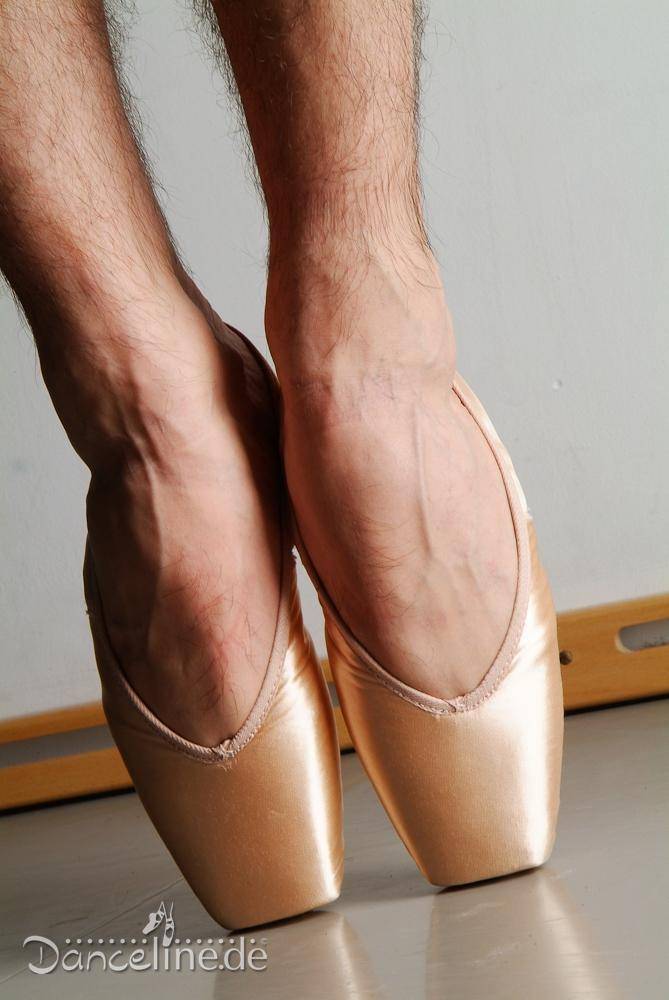 Eine Nahaufnahme der Füße einer Tänzerin, die Spitzenschuhe Merlet Pulsion von Dance Line trägt, die für Ballett entworfen wurden. Die Schuhe sind rosa und die Tänzerin steht auf den Zehenspitzen, wodurch ihre Wadenmuskeln betont werden. Im Hintergrund ist der untere Teil der Ballettstange zu sehen. In der unteren linken Ecke steht der Text „Danceline.de“.