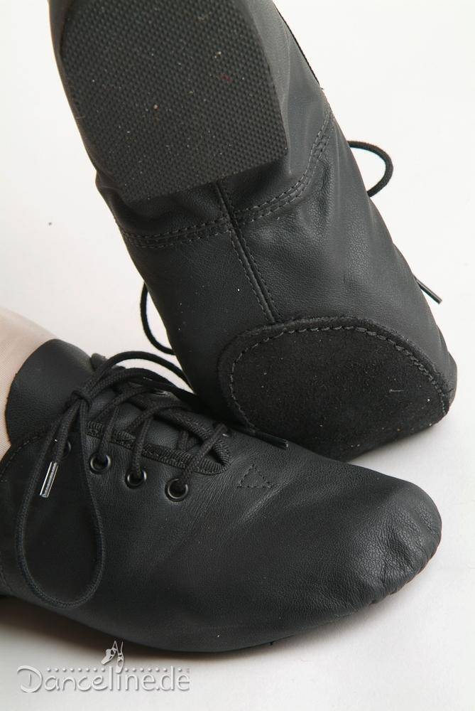 Nahaufnahme eines Paars schwarzer Jazzschuhe SoDanca JZE09A Jazzme, die von einer Person getragen werden, die auf einer hellen Oberfläche steht. Die gut sitzenden Schuhe zeigen die Sohle und den oberen Teil, wobei in der unteren linken Ecke das Logo „Dancelines.de“ sichtbar ist.