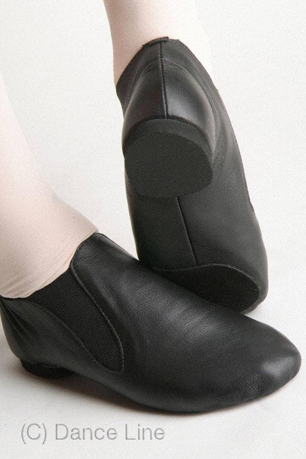 Nahaufnahme einer Person, die schwarze Jazzschuhe SoDanca JZE40A Elasto mit elastischen Seiten trägt. Die Chromledersohle ist von der Seite und von unten zu sehen, was die glatte Textur und Flexibilität hervorhebt. Die Person steht auf einer hellen Oberfläche. (C) SoDanca.