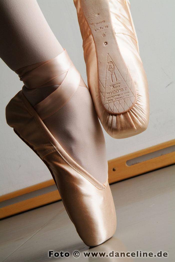 Nahaufnahme der Füße einer Balletttänzerin in rosa Spitzenschuhen von Bloch S0141LV Serene, in einer klassischen Ballettpose. Im Hintergrund sind eine hölzerne Ballettstange und ein Teil einer weißen Wand zu sehen. Die Schuhe haben sichtbare Texte und Markierungen auf den Sohlen, die ihre breite Standfläche zeigen.