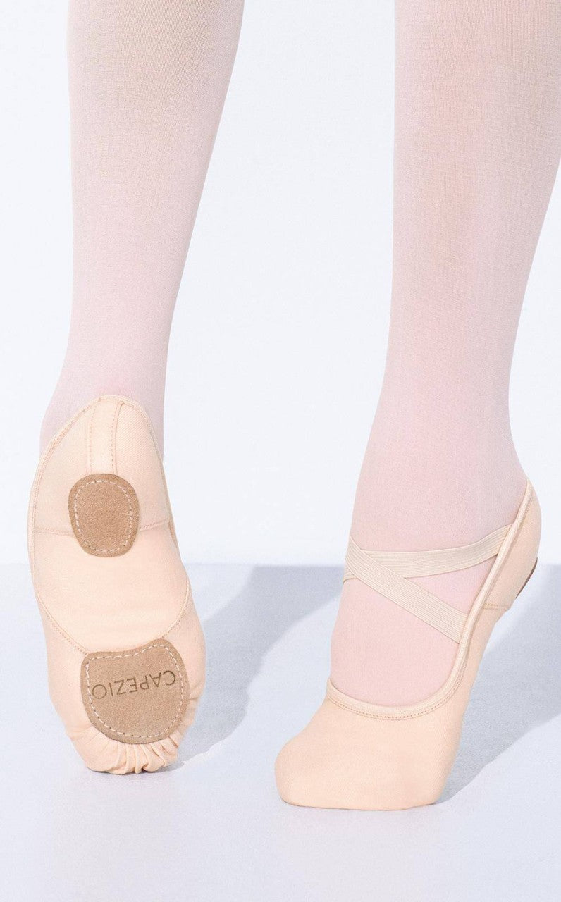 Nahaufnahme der Unterschenkel und Füße einer Tänzerin, die hellrosa Ballettschuhe Capezio 2037W Hanami und Strumpfhosen trägt. Der linke Fuß steht flach auf dem Boden und zeigt die geteilte Chromledersohle mit der Aufschrift „Capezio“, während der rechte Fuß leicht angehoben ist und elastische Bi-Stretchleinen-Riemen zeigt.