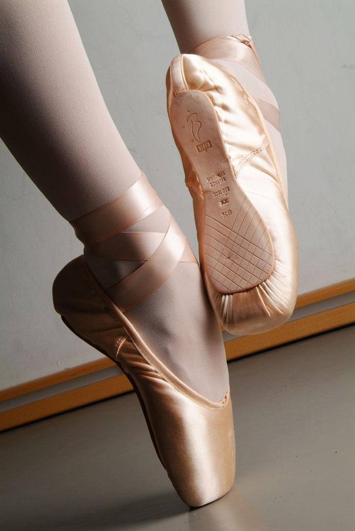 Eine Nahaufnahme der Füße einer Balletttänzerin in rosa Spitzenschuhen Bloch S0160S Balance European Strong. Die fortgeschrittenere Tänzerin balanciert auf den Spitzen und zeigt die Abnutzung der harten Sohle. Die Bänder sind ordentlich um die Knöchel gebunden und die Tänzerin trägt helle Ballettstrumpfhosen.