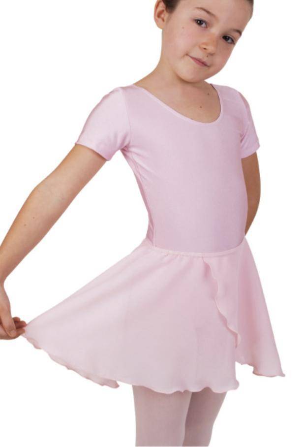Ein junges Mädchen steht in einem rosa Ballett-Outfit und hält die Ränder ihres Rocks in Wickeloptik Danceries Z39 Lilly von Danceries seitlich nach außen. Sie scheint anmutig zu posieren, mit einem leichten Lächeln und dem Kopf zur Seite geneigt. Sie hat kurze Haare und sieht in ihrem zarten Ensemble aus Georgette-Stoff so aus, als wäre sie bereit für eine Tanzvorführung oder eine Übung.