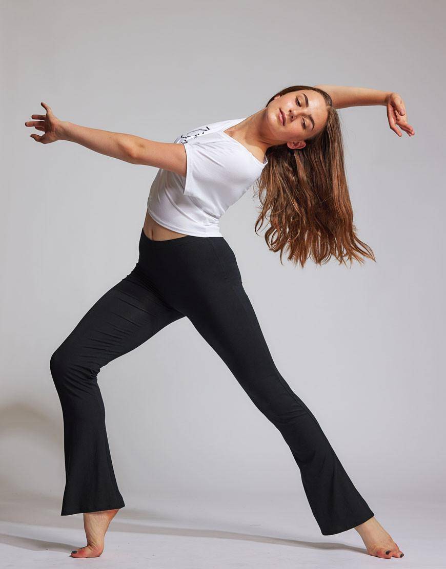 In einem Studio führt Luna eine Tanzbewegung aus, ein Bein ist nach hinten ausgestreckt und das andere leicht gebeugt. Ihr langes braunes Haar weht, während sie auf ihren Zehenspitzen steht und die schwarze Jazzhose TempsDanse Luna und ein weißes bauchfreies Top trägt. Mit anmutig ausgestreckten Armen verkörpert sie die Essenz von Temps Danse.