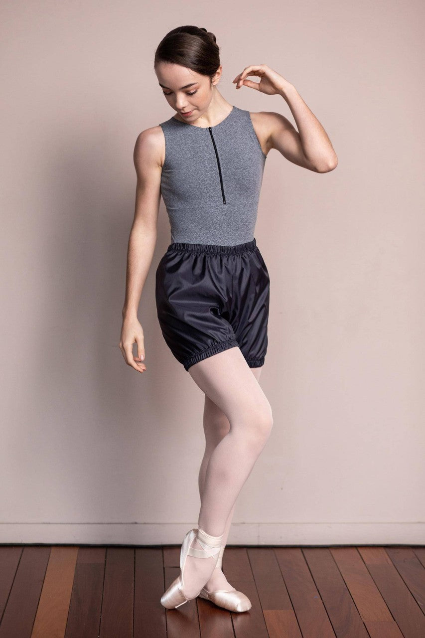 Eine Balletttänzerin in einem grauen ärmellosen Trikot und schwarzen Intermezzo Wärmeshorts 5271 Sauna steht auf Spitze auf einem Holzboden. Sie trägt weiße Strumpfhosen und Ballettschuhe, ihr rechter Arm ist am Ellbogen angewinkelt und ihr linker Arm nach unten ausgestreckt. Der Hintergrund ist eine schlichte beige Wand.