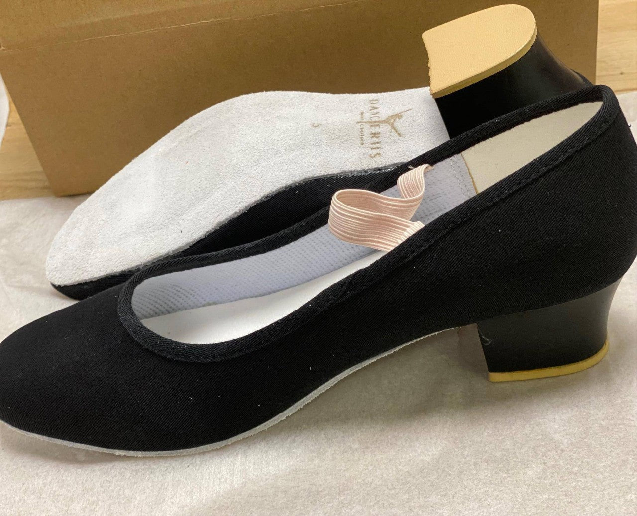 Ein Paar Charakterschuhe Danceries Z66 in Schwarz mit niedrigem Absatz und elastischem Riemen oben. Diese exquisiten Charakter-Tanzschuhe von Danceries, empfohlen von der Royal Academy of Dance, werden nebeneinander ausgestellt – einer zeigt die Draufsicht und der andere hebt Sohle und Absatz hervor. Im Hintergrund ist ein Karton zu sehen.