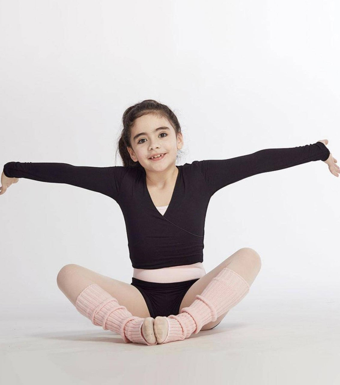 Ein junges Mädchen sitzt mit überkreuzten Beinen und ausgestreckten Armen vor einem einfarbigen Hintergrund auf dem Boden. Sie lächelt und trägt einen schwarzen Turnanzug, hellrosa Beinwärmer, um ihre Muskeln warm zu halten, und ein pfirsichfarbenes Temps Danse Wickeltop TempsDanse Vidy Junior aus weicher Viskose.