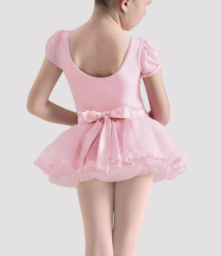 Ein kleines Kind trägt eine Kindertütülette Bloch CR4061Rose von Bloch, mit kurzen Ärmeln und einem Tüll-Tutu, von hinten betrachtet. Das Tutu weist Rüschenlagen auf und ist an der Taille mit einer großen Satinschleife verziert. Das Kind hat kurze Haare und steht vor einem schlichten, hellgrauen Hintergrund.