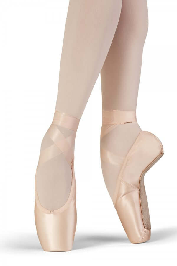 Die Füße einer Balletttänzerin tragen hellrosa Spitzenschuhe Bloch S0161L Grace und beige Strumpfhosen. Ein Fuß steht flach auf dem Boden, während der andere gestreckt und angehoben ist, was zeigt, wie die fortgeschrittene Tänzerin auf den Zehenspitzen balanciert.