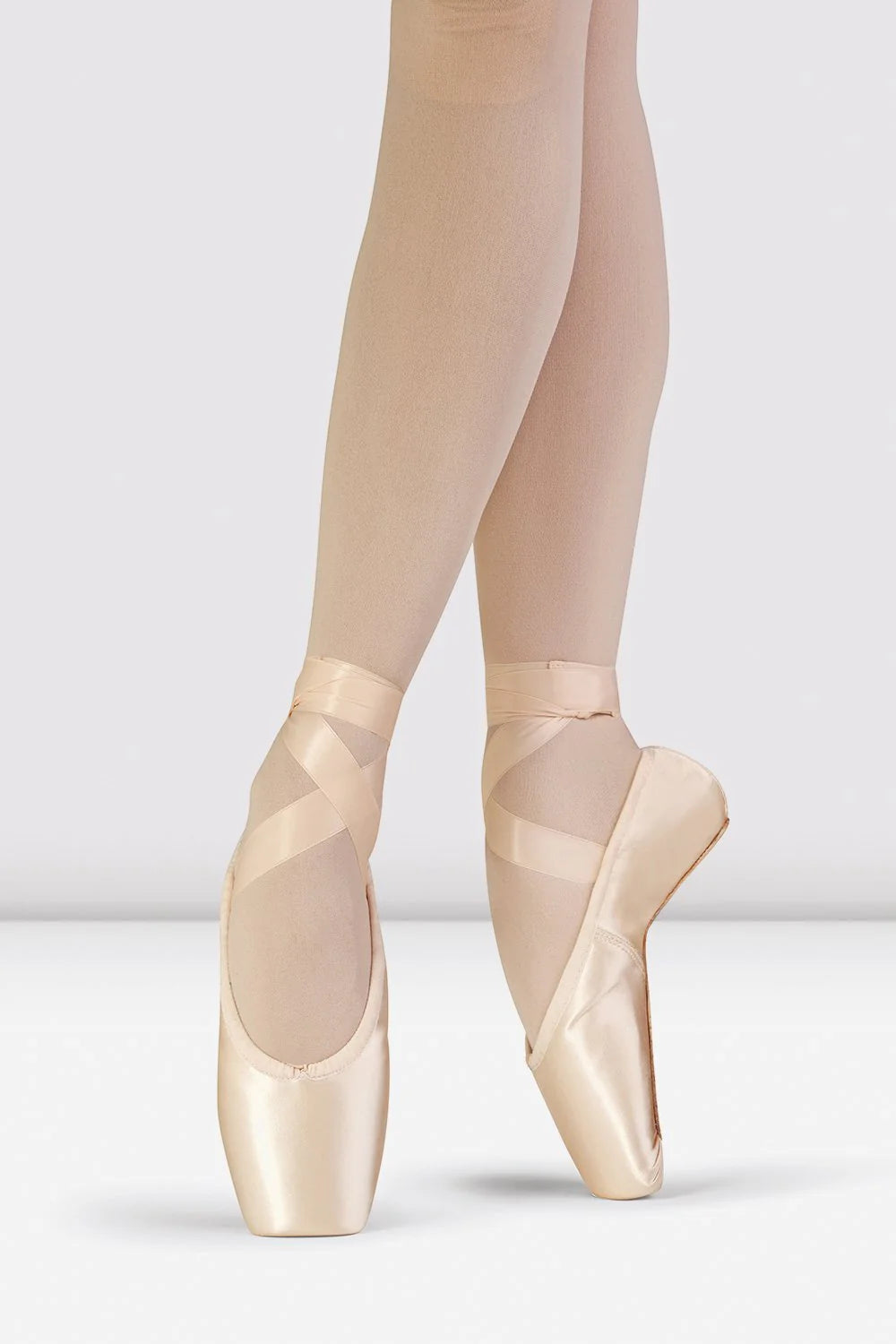 Nahaufnahme einer Ballerina auf Spitze, die Bloch Spitzenschuhe S0175L Synthesis und beige Strumpfhosen trägt, wobei ihre Füße anmutig vor einem hellgrauen Hintergrund positioniert sind.