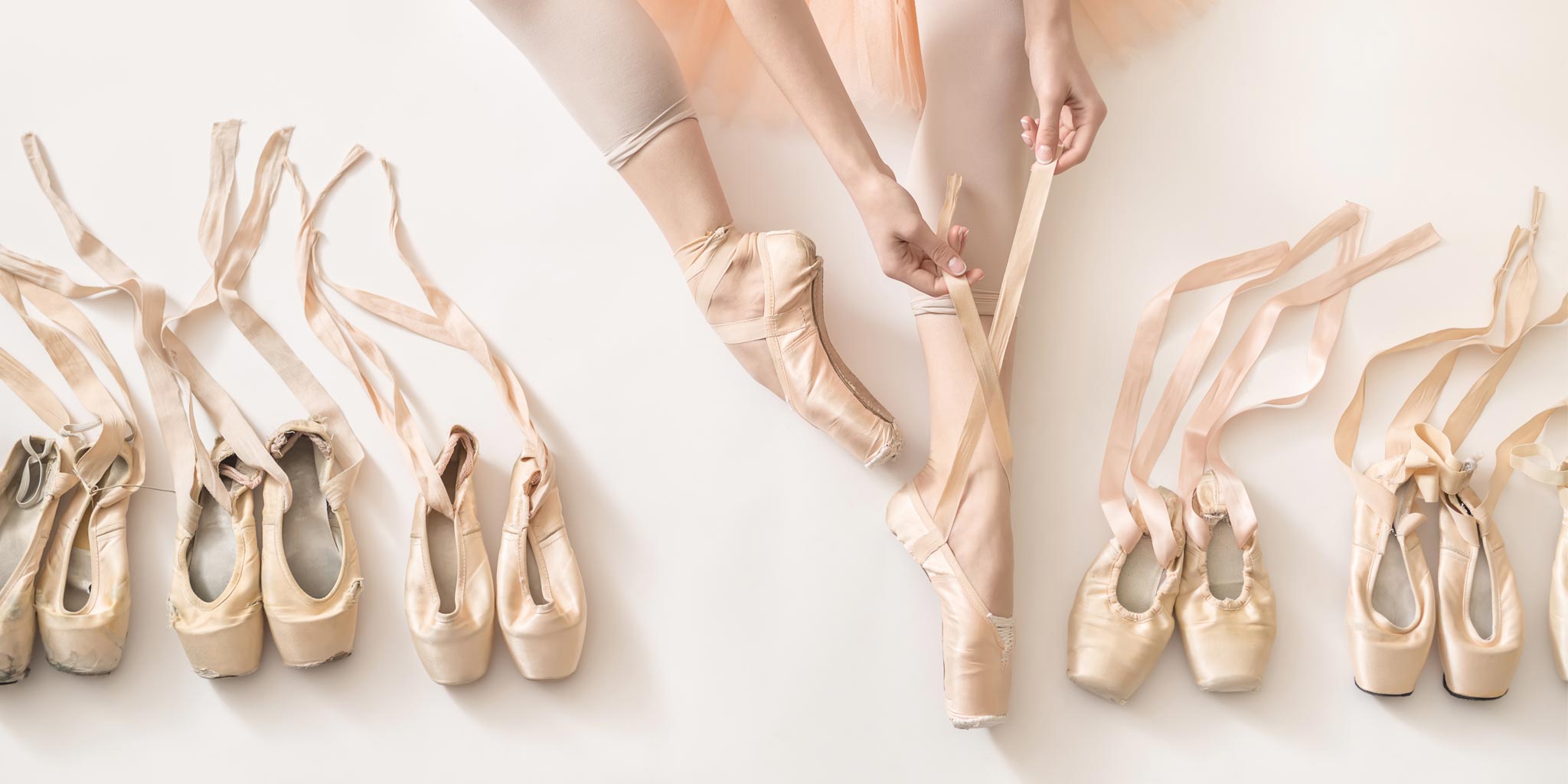 Viele gebrauchte Spitzenschuhe mit Bändern. In der Mitte sind die Beine einer Ballerina. Sie trägt Spitzenschuhe und bindet einen Spitzenschuh gerade.