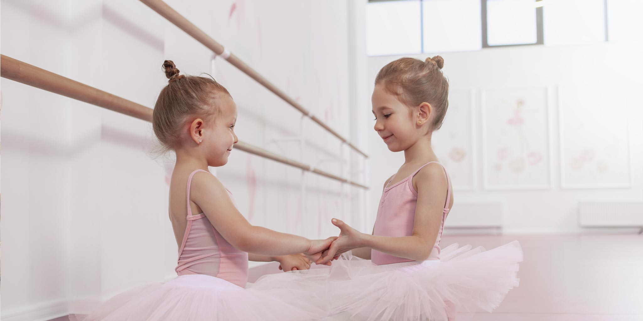 Zei kleine Ballettmädchen in einem Ballettsaal. Sie haben rosa Ballettkleider an mit Tüllrock und halen sich an den Händen. Im Hintergrund ist eine Ballettstange zu sehen.