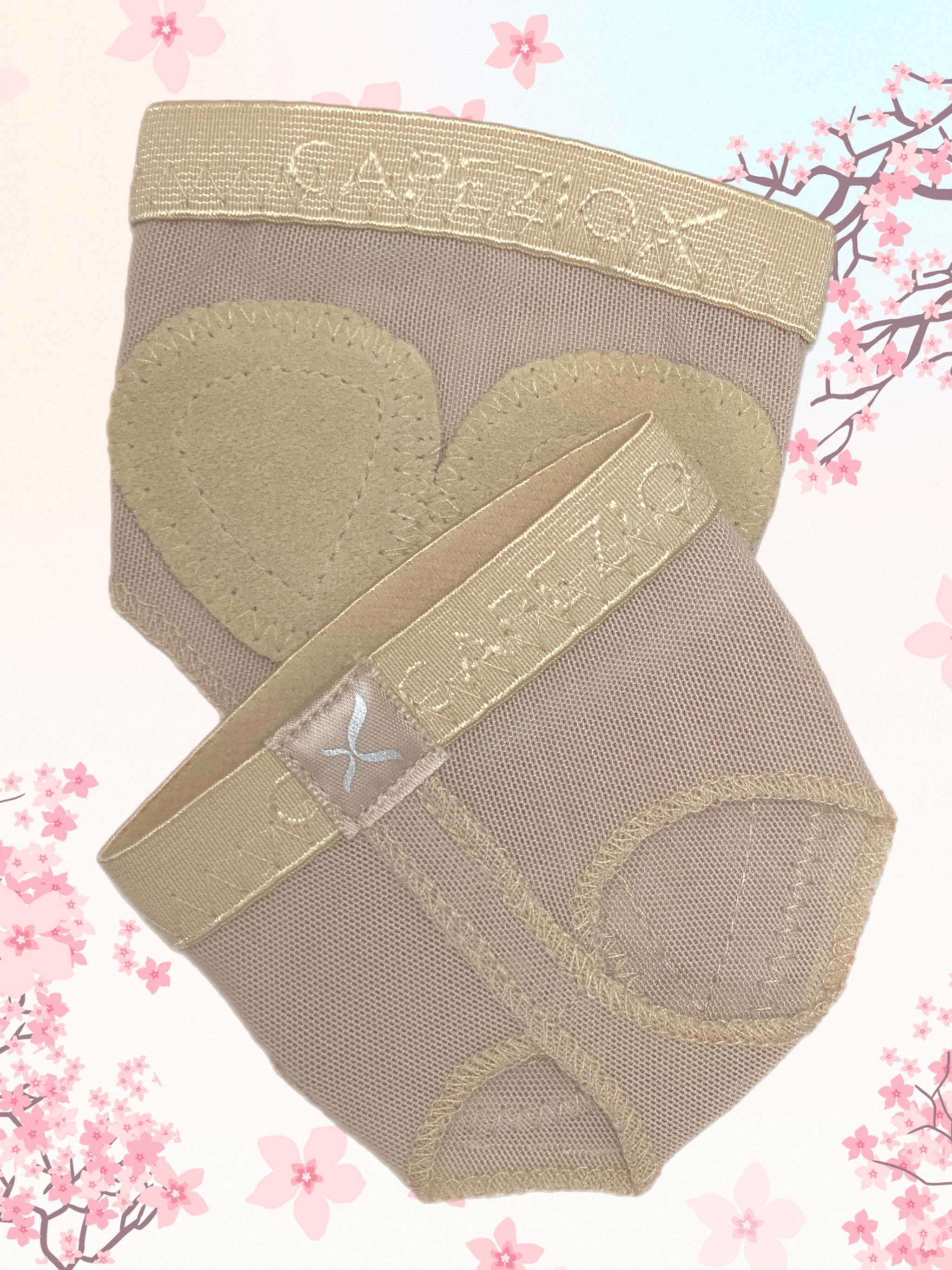 Ein Paar Ballenschutz Capezio H07B FootUndeez in Beige, mit herzförmigen Polstern aus Spandex-Material vor einem Hintergrund aus hellrosa Kirschblüten-Illustrationen. Ausgestattet mit einem elastischen Riemen ist der Markenname „Capezio“ sowohl auf dem Bund als auch auf dem Riemen deutlich zu erkennen.