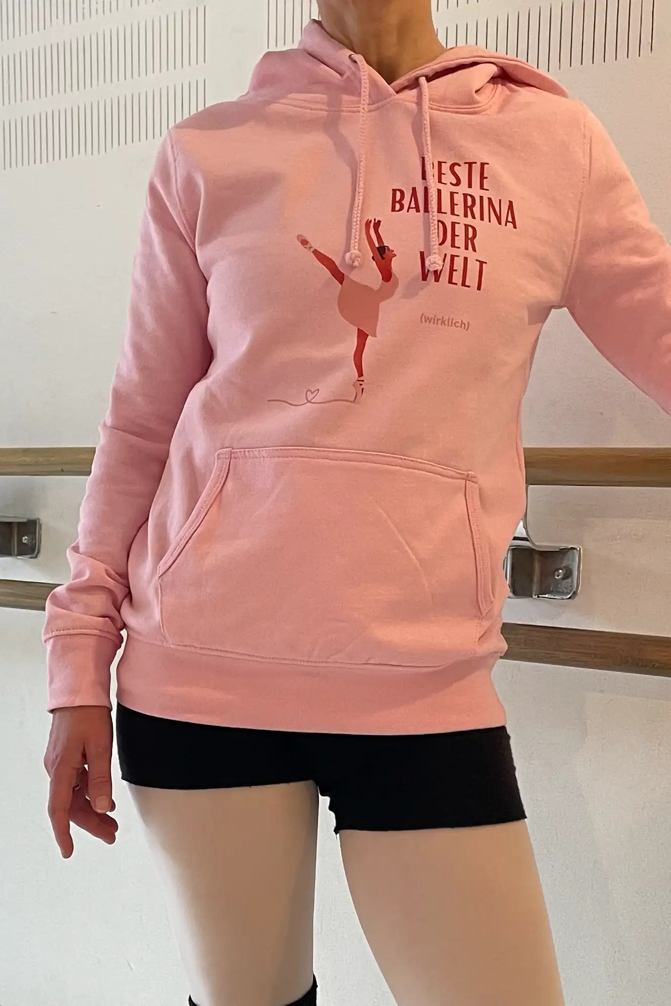 Eine Person trägt den Dance Line Ballett Hoodie Danceline Beste Ballerina, der eine Ballerina-Illustration und den deutschen Satz „Beste Ballerina der Welt“ zeigt. Sie befindet sich in einem Tanzstudio, steht neben einer Ballettstange und trägt schwarze Tanzshorts.