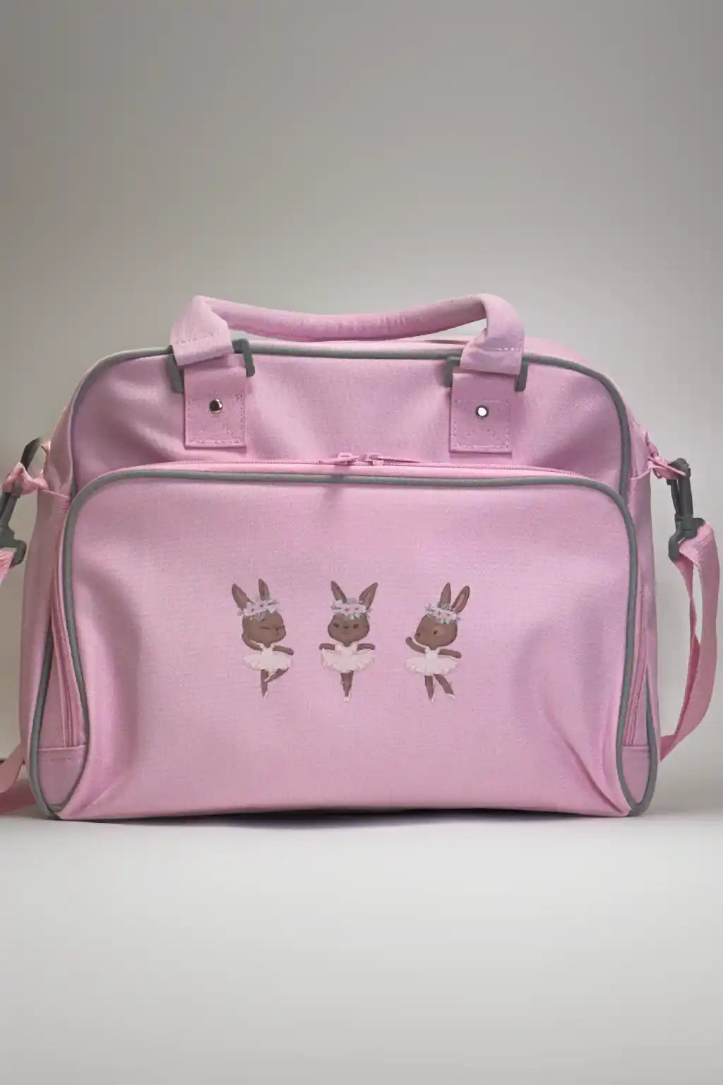 Die Kinder-Balletttasche Danceline Ballet Bears von Danceline ist eine rosa Tragetasche mit zwei Tragegriffen, einem abnehmbaren Schultergurt und drei gestickten Hasen in Tutus auf der Vordertasche.