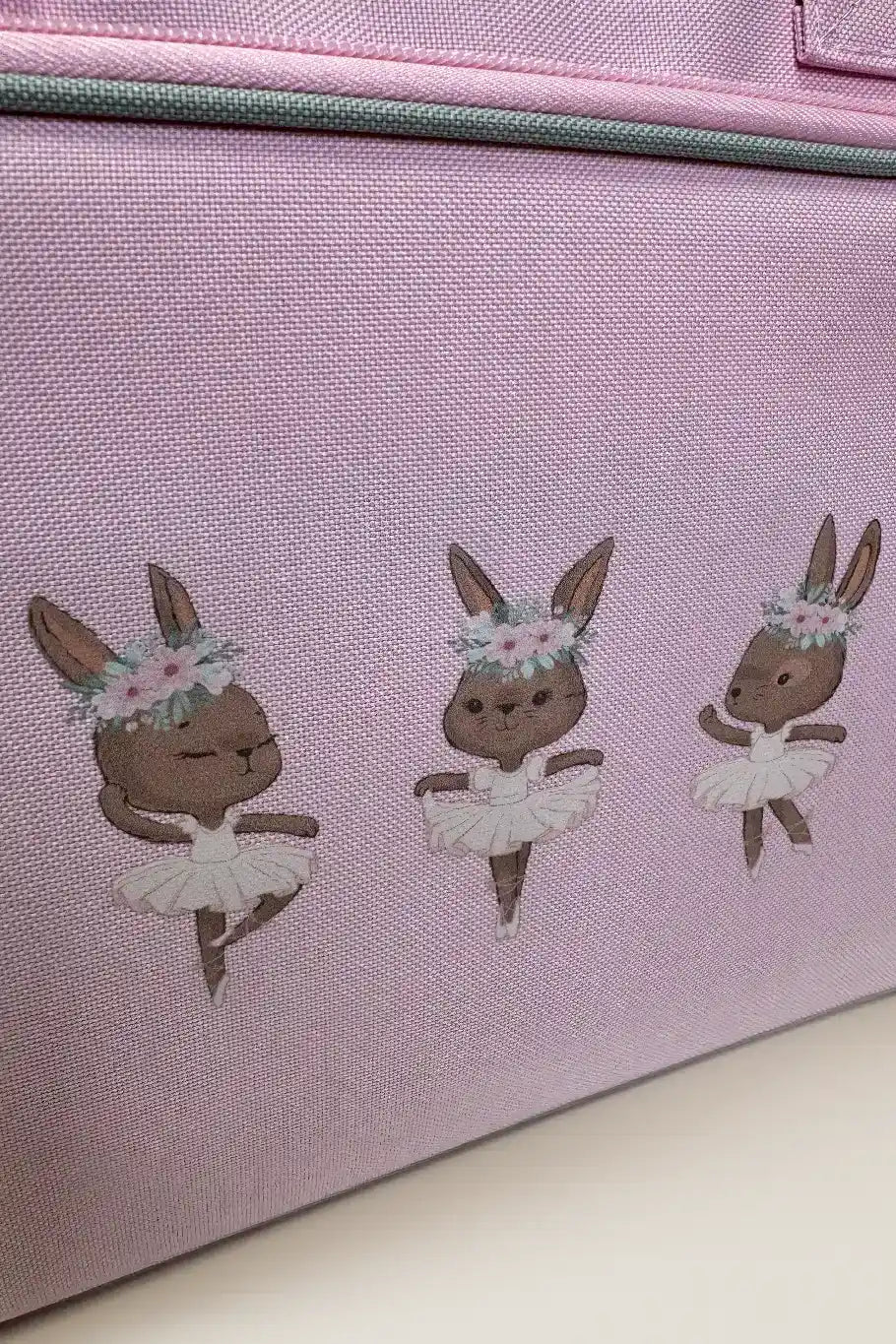 Die Kinder Balletttasche Danceline Ballet Bears von Danceline besteht aus lavendelfarbenem Stoff und ist mit Bildern von drei balletttanzenden Häschen verziert. Jedes Häschen trägt ein weißes Tutu und einen Blumenkopfschmuck und zeigt verschiedene Ballettposen vor einem zartrosa Hintergrund.