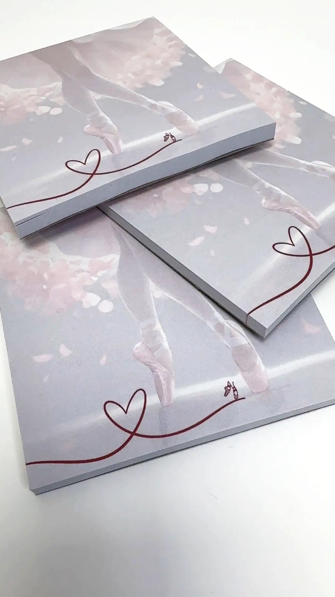 Piles of Block Danceline-Notizbücher „Dancing around“ von Dance Line mit einem Ballettthema in sanften Rosa- und Weißtönen. Das Coverdesign hebt Ballettschuhe und eine durchgehende rote Linie hervor, die in der Ecke eine Herzform bildet.