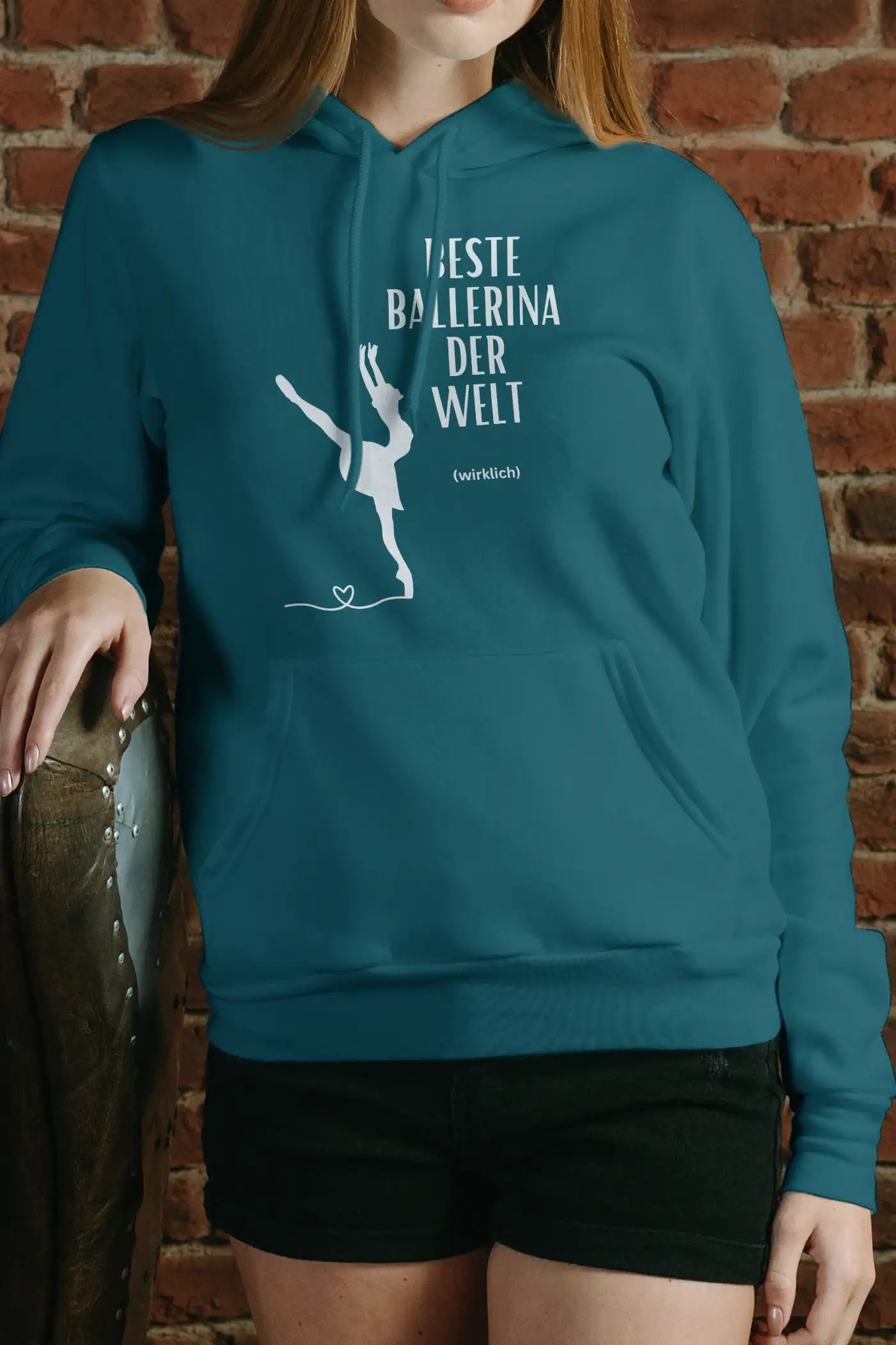 Eine Person, die den Dance Line Ballett Hoodie „Danceline Beste Ballerina“ trägt, der die Silhouette einer Balletttänzerin und den deutschen Text „Beste Ballerina der Welt (wirklich)“ zeigt, steht vor einer Backsteinwand und ruht mit einer Hand auf einem Ledersessel.