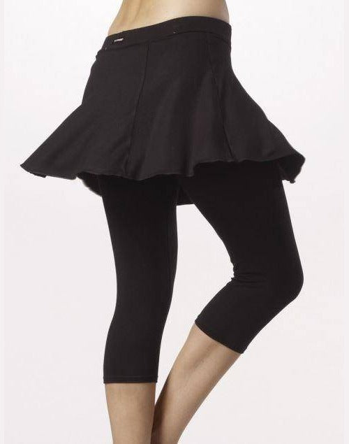 Person, die nur ihren Unterkörper zeigt und einen schwarzen Skaterrock über der bequemen, elastischen Viskose-Caprihose Temps Danse Hose TempsDanse Zazou trägt. Der Rock ist am Saum ausgestellt und die Leggings sind knapp unter dem Knie abgeschnitten. Der Hintergrund ist schlicht und hell.