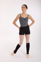 Eine Balletttänzerin, elegant gekleidet in einem grauen Trikot, schwarzen Shorts und 50 cm langen Beinwärmern „Stulpen“ von Intermezzo (Modellnummer 2034 Danae), steht auf Spitzenschuhen und blickt zur Seite vor einem schlichten weißen Hintergrund.