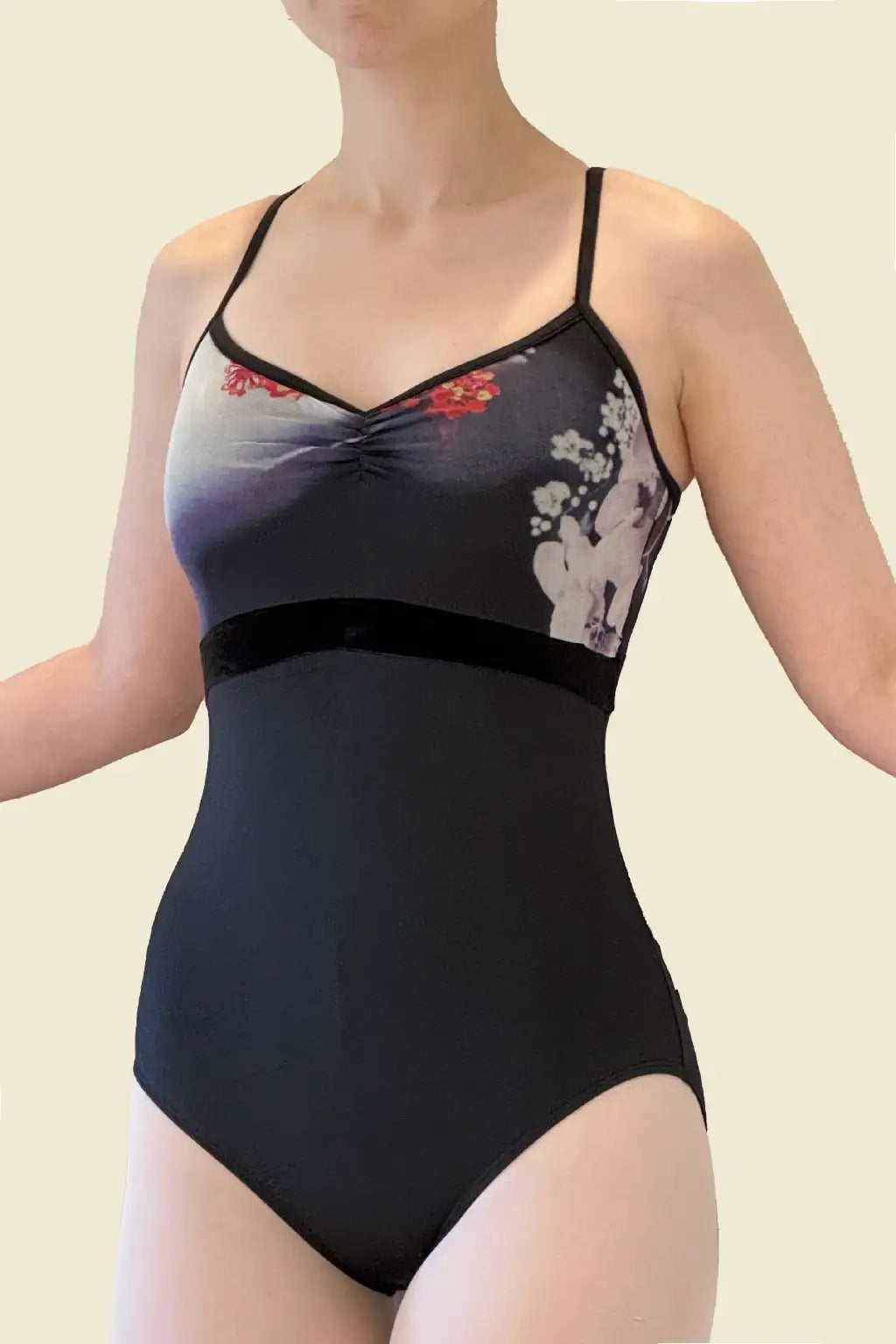 Eine Person trägt den Ballettanzug Damen Sansha Gillian 0068M von Dance Line, ein schwarzes einteiliges Design mit dünnen Trägern und einem Blumen- und Schmetterlingsmuster auf der Oberseite, vor einem schlichten hellen Hintergrund.