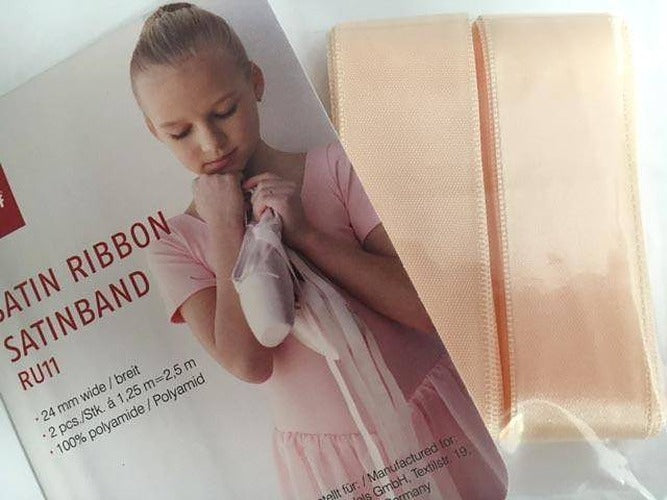 Eine Rumpf Satinbänder Rumpf RU11 Produktverpackung mit einem Bild eines jungen Mädchens in einem Ballett-Outfit, das rosa Spitzenschuhe hält. Die Verpackung enthält breite hellrosa Satinbänder, die als 2,4 cm breit und 2,5 Meter lang beschrieben werden und aus Seide und Polyamid bestehen.